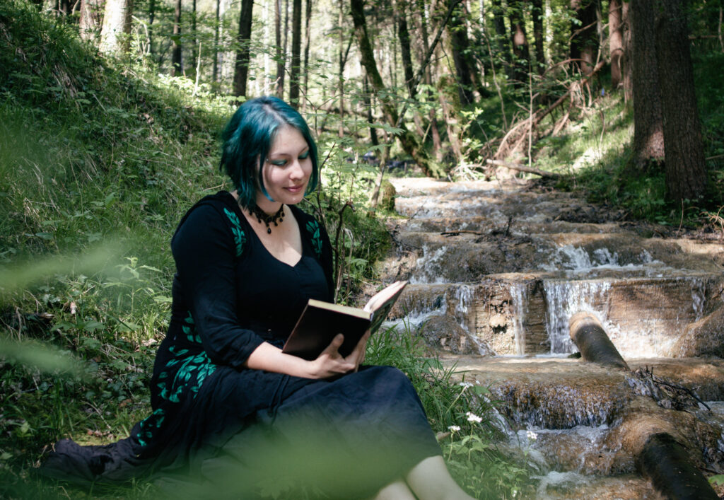 Die Autorin sitzt lesend im Wald. Sie ist umgeben von grünen Pflanzen. Neben ihr plätschert ein Gebirgsbach.